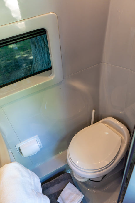 Inside a compact RV rental bathroom: a white toilet, toilet paper holder, and window with trees visible. Towels on the floor.