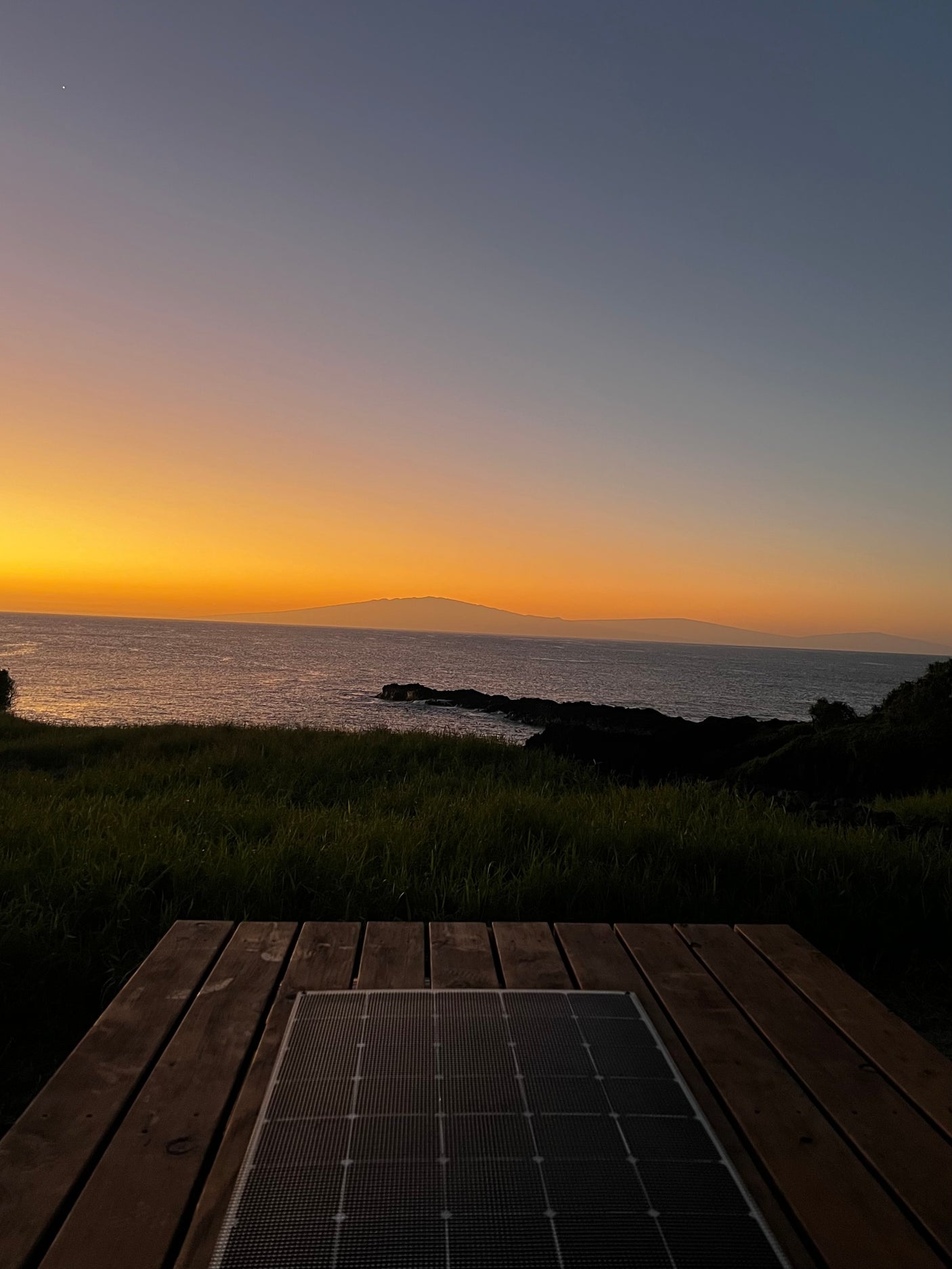 A vibrant orange and blue sunset over the ocean, with distant land. A solar panel on a wooden deck suggests an RV rental setup.
