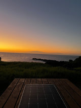 A vibrant orange and blue sunset over the ocean, with distant land. A solar panel on a wooden deck suggests an RV rental setup.