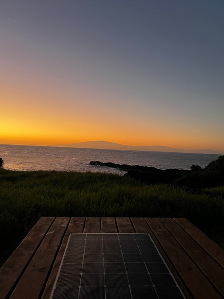 A vibrant orange and blue sunset over the ocean, with distant land. A solar panel on a wooden deck suggests an RV rental setup.