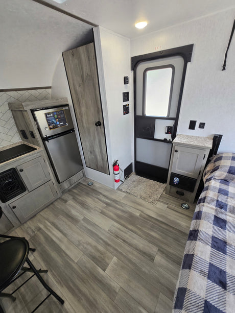 Modern rv rental interior showcasing a kitchenette, fridge, entry door, fire extinguisher, and bed with plaid blanket. Compact design.