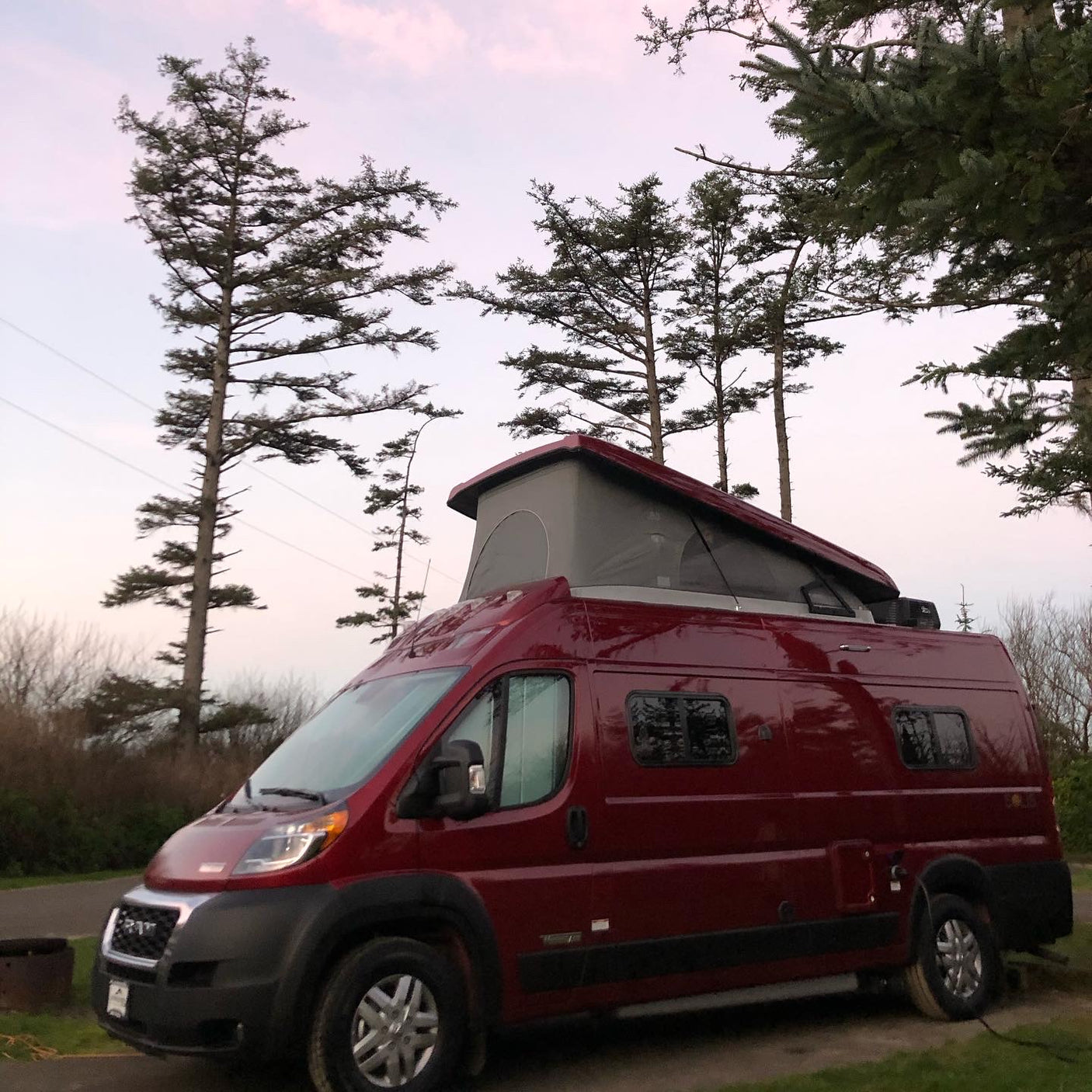 Red pop-top RV rental parked at a serene campsite among tall trees at dusk. Perfect for outdoor adventure.