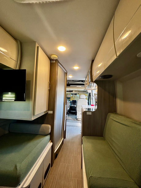 An RV rental interior shows a narrow hallway leading to the driver's cabin, flanked by green upholstered benches, a kitchen, and overhead cabinets.