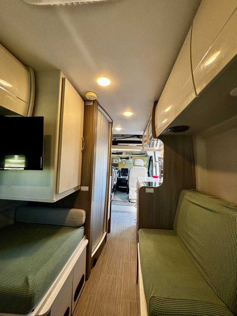 An RV rental interior shows a narrow hallway leading to the driver's cabin, flanked by green upholstered benches, a kitchen, and overhead cabinets. — photo 15
