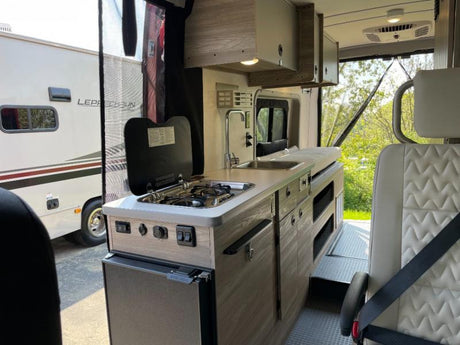 Explore mobile living! This rv rental interior boasts a compact kitchen with a stove, sink, and fridge, plus a passenger seat.