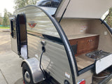 This RV rental teardrop camper features an open side door to a cozy bed and a rear outdoor kitchen with stove and sink.