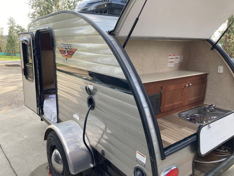 This RV rental teardrop camper features an open side door to a cozy bed and a rear outdoor kitchen with stove and sink.
