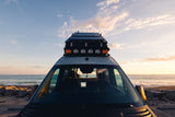 An RV rental camper van with rooftop solar panels & lights overlooks the ocean at golden hour.