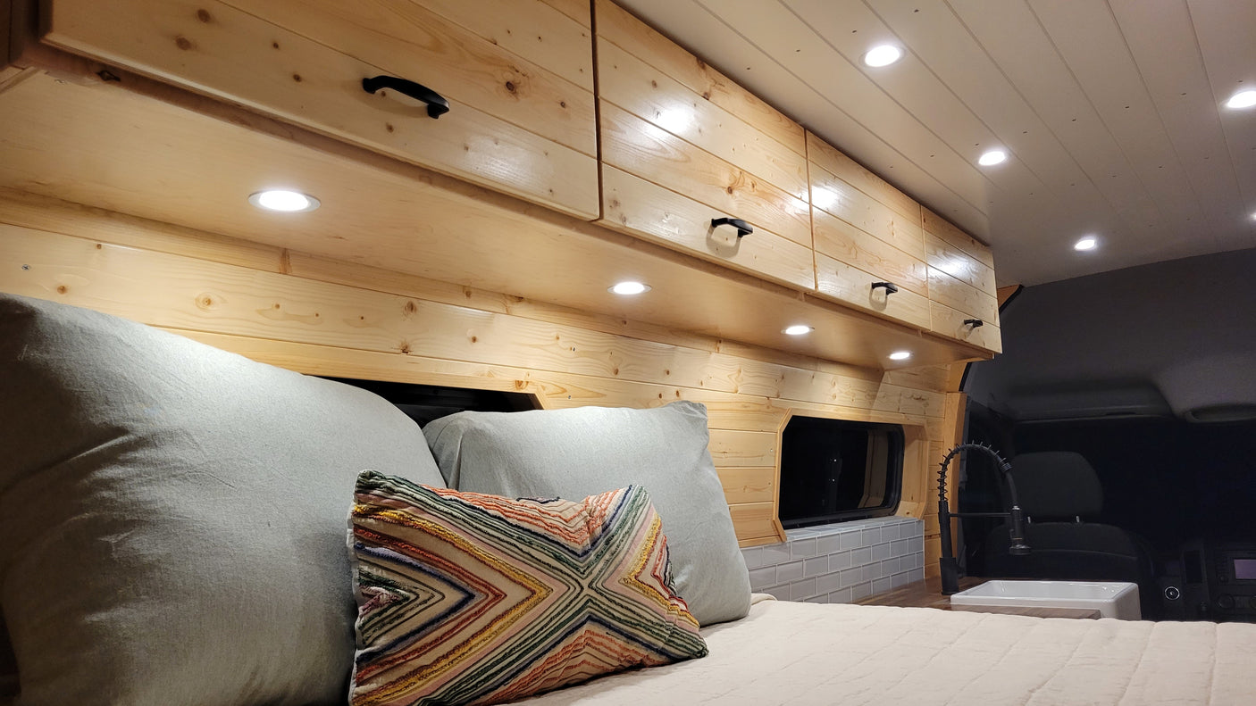 Inside a bright RV rental: wooden walls, overhead cabinets, a cozy bed, and a compact sink. Ideal mobile living.