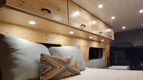 Inside a bright RV rental: wooden walls, overhead cabinets, a cozy bed, and a compact sink. Ideal mobile living.