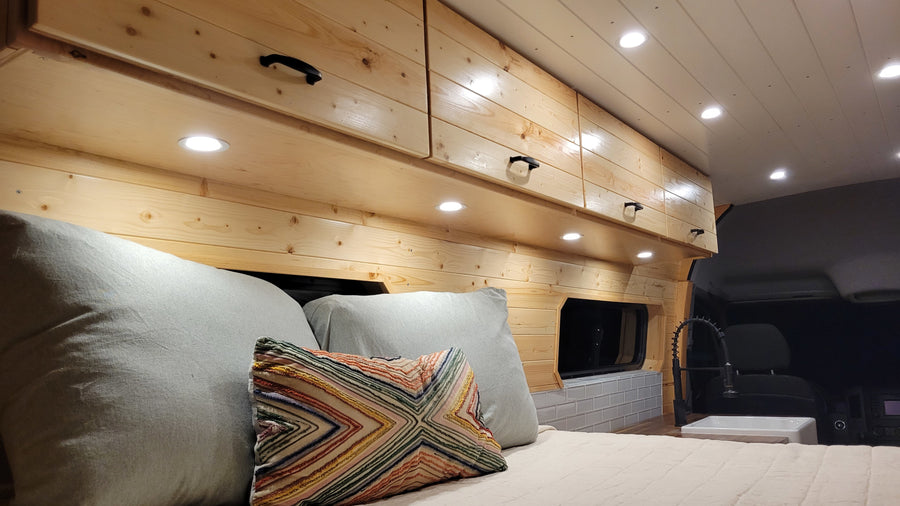Inside a bright RV rental: wooden walls, overhead cabinets, a cozy bed, and a compact sink. Ideal mobile living.