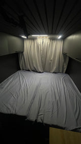 Cozy RV rental sleeping area: bed with grey sheets, privacy curtains, & LED lights in a dark, compact interior.