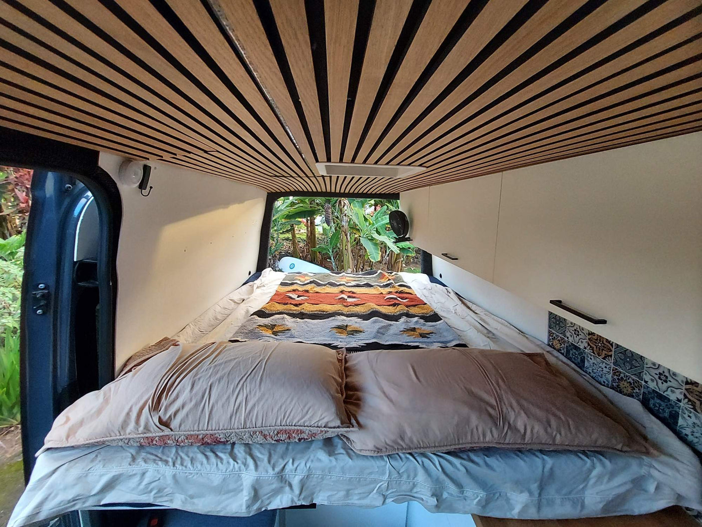 Comfortable camper van interior for your next rv rental adventure. Features a cozy bed, unique slatted ceiling & lush nature view.