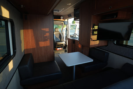 Bright rv rental interior showing dining, kitchenette, TV, and driver's cabin, illuminated by sunlight.