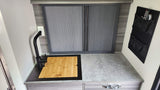 Compact RV rental kitchen counter with sleek black faucet, built-in sink/cutting board, and tambour storage unit.