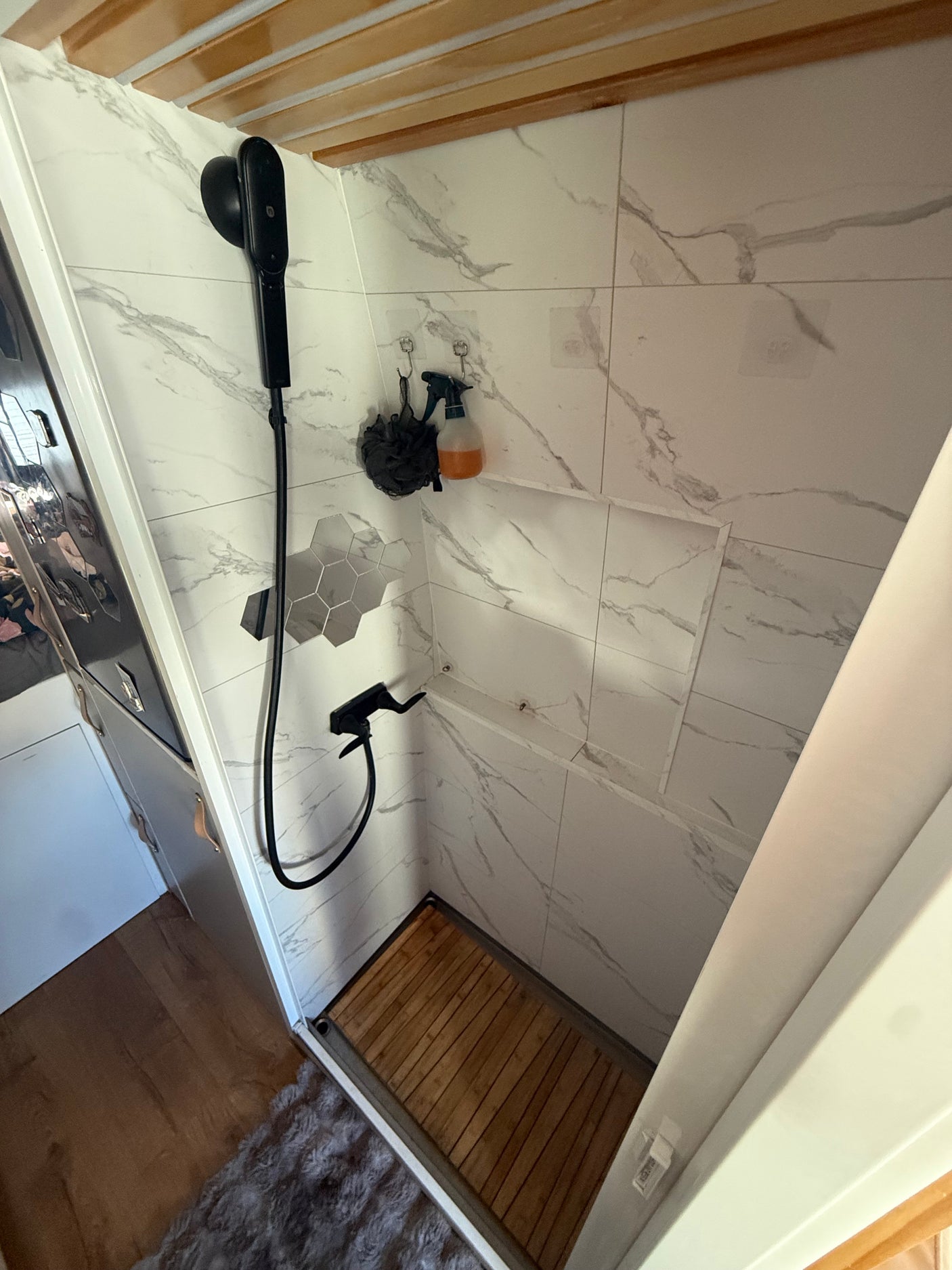 This modern, compact RV rental shower features white marble-look tiles, black fixtures, and a wooden slatted floor.