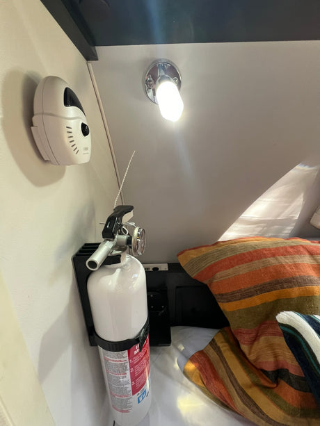 An RV rental features a mounted fire extinguisher, a white detector, and striped bed pillows, offering essential safety and compact living.