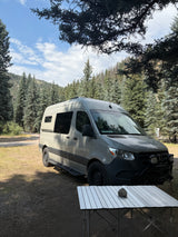 A light-colored Sprinter camper van (RV rental) is parked in a pine forest, with a metal table in the foreground.