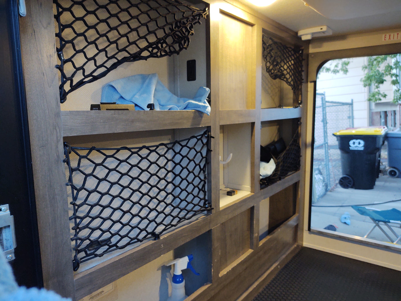 RV rental interior: organized wooden shelves with black cargo nets, holding a blue towel. View outside door.