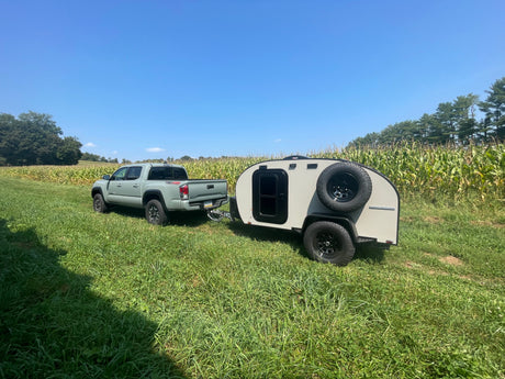 Green Toyota Tacoma towing a teardrop RV rental in a grassy field next to a cornfield under a clear blue sky.