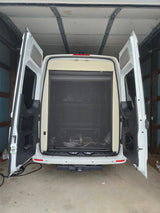 White camper van (RV rental) in a garage, rear doors open. A black mesh screen covers the interior, with a hitch below.