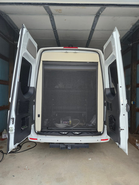 White camper van (RV rental) in a garage, rear doors open. A black mesh screen covers the interior, with a hitch below.