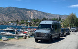 Gray Mercedes-Benz Sprinter RV rental parked by a bustling lake marina, surrounded by scenic mountains and lakeside homes.