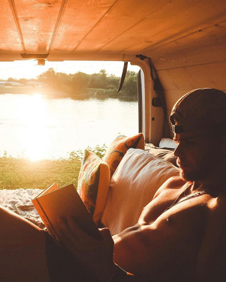 A man reads a book inside an RV rental, lit by golden sunset light over a river. Perfect for travel & relaxation.