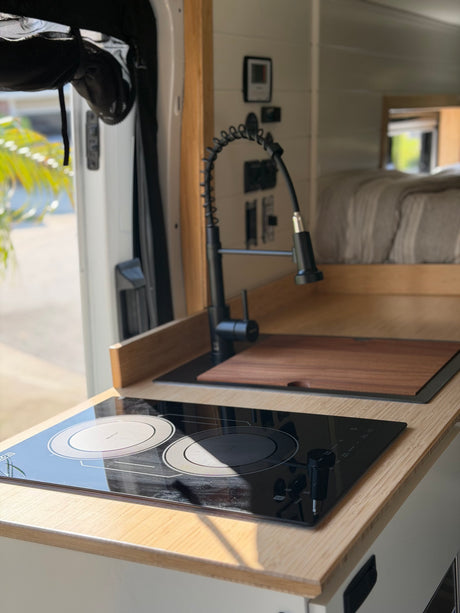 Stylish rv rental kitchen area: two-burner induction cooktop, black faucet, and sink with wooden cutting board.