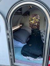 Inside this cozy RV rental, discover a comfortable bed, built-in shelf with outlets, and a unique arched window view.