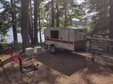 Compact teardrop RV rental at a scenic lakeside campsite under tall trees, ideal for outdoor adventures.