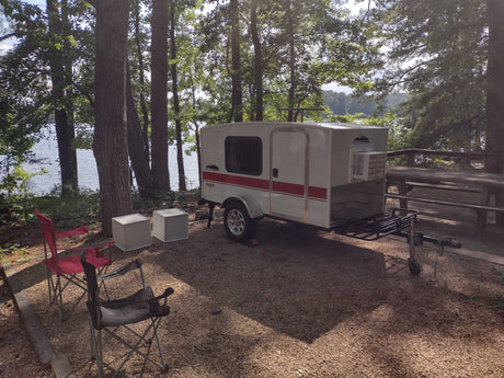 Compact teardrop RV rental at a scenic lakeside campsite under tall trees, ideal for outdoor adventures.