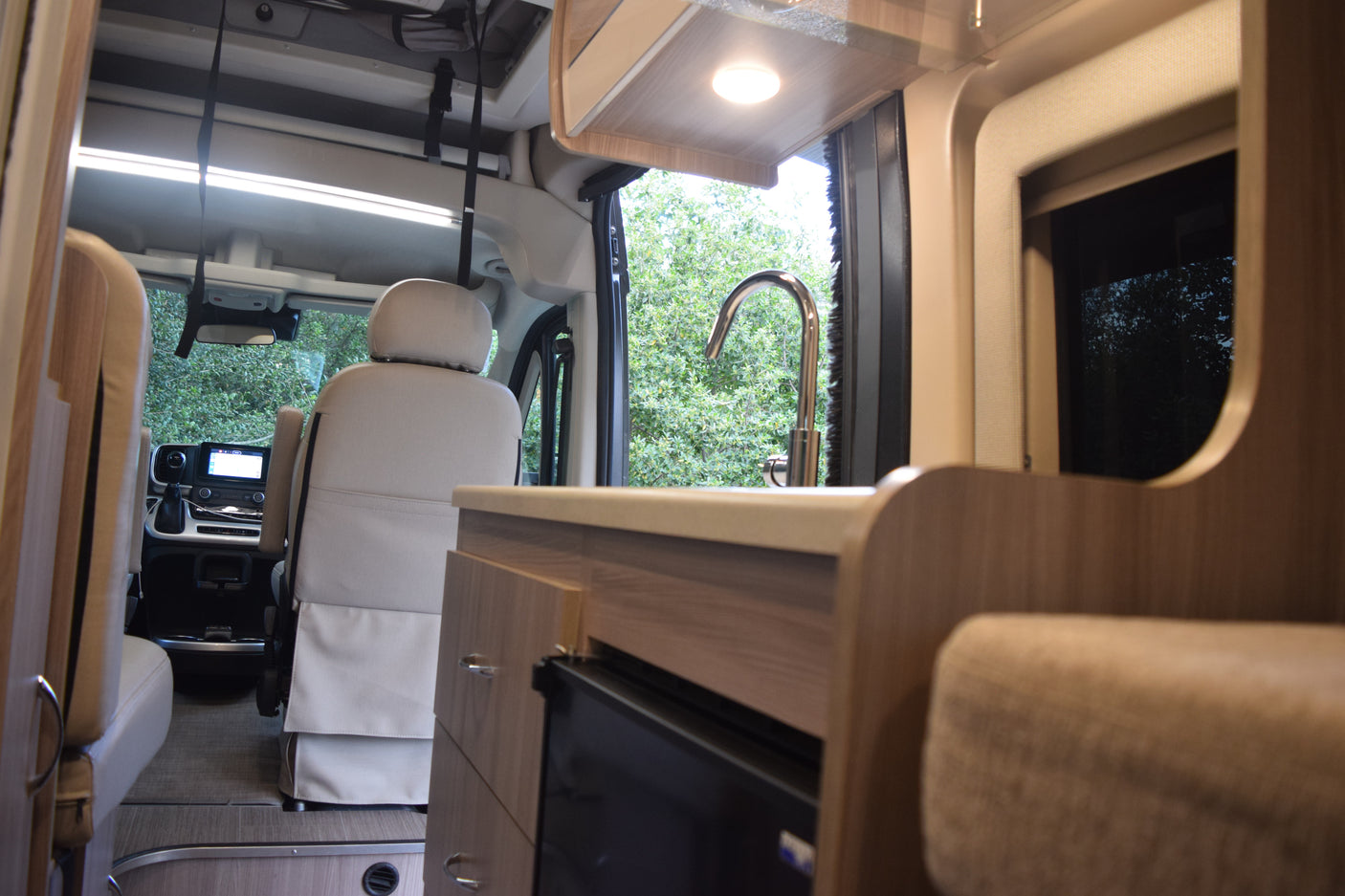 Modern rv rental interior showing front cab, kitchen with sink & faucet, integrated fridge, and light wood cabinetry. Green views outside.
