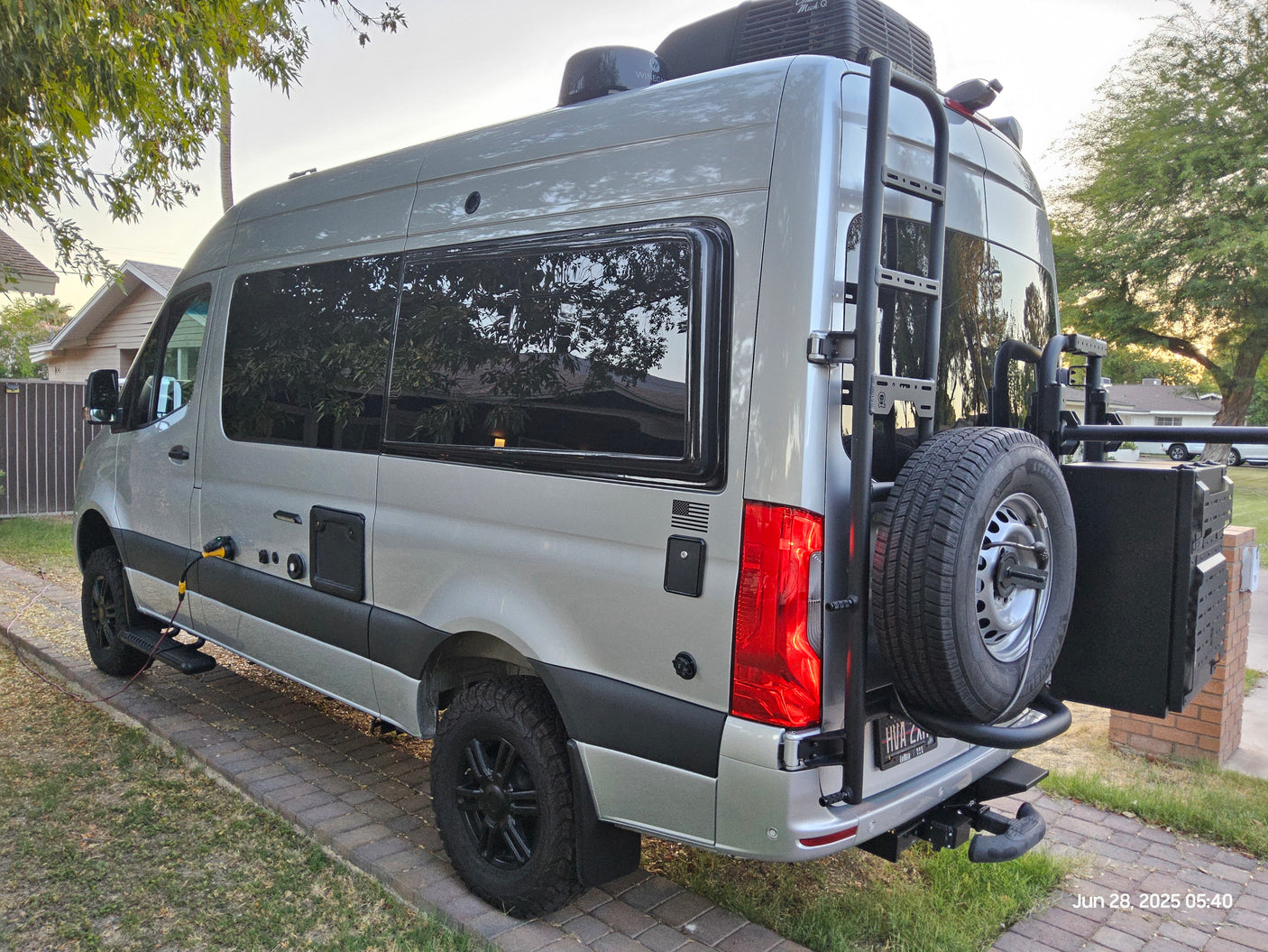 A silver camper van, customized with roof AC, rear ladder & spare, parked on a brick driveway. Great for an RV rental trip.
