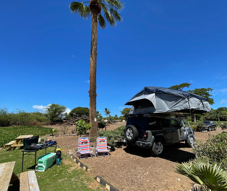 Experience outdoor adventure! This RV rental-style Jeep with a rooftop tent is set up at a sunny campsite with chairs & cooking gear.