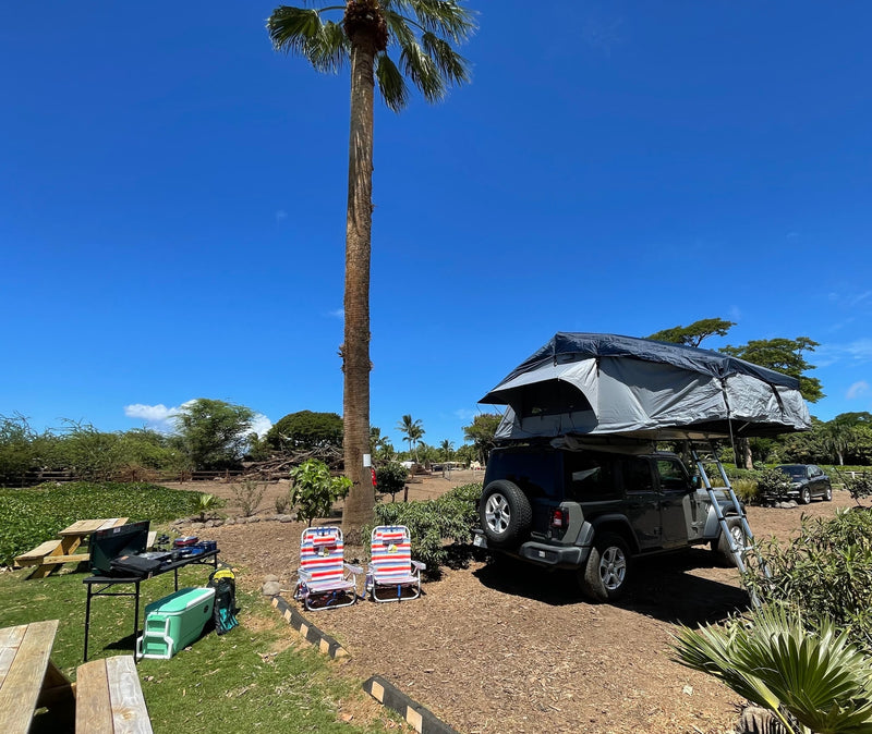  parked for camping under a bright blue sky.|Experience outdoor adventure! This RV rental-style Jeep with a rooftop tent is set up at a sunny campsite with chairs & cooking gear.|This Jeep with a rooftop tent is an ideal RV rental for exploring Hawaii's scenic mountains and lush campsites.|Hawaii RV rental: grey Jeep Wrangler with rooftop tent at a sunny campsite (site 11).|Jeep Wrangler with a rooftop tent and ladder — photo 2