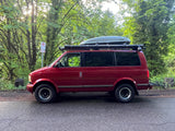 Red van, equipped with a roof cargo box and rear rack, an ideal rv rental for outdoor adventures, parked by a sunny forest.