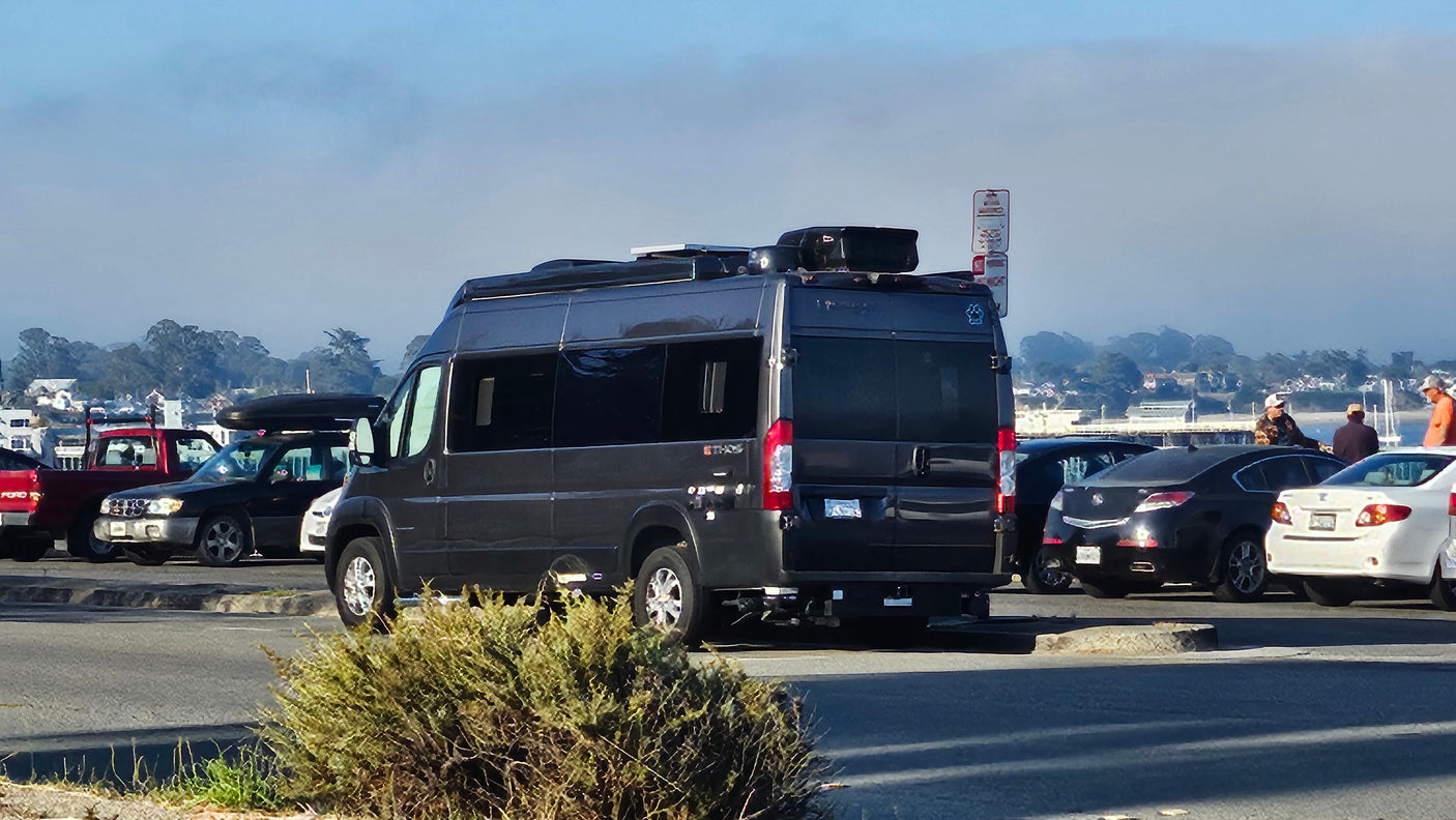 A dark grey camper van, like an RV rental, with roof storage, parked in a sunny lot with other vehicles and people.