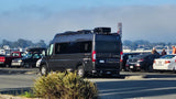 A dark grey camper van, like an RV rental, with roof storage, parked in a sunny lot with other vehicles and people.