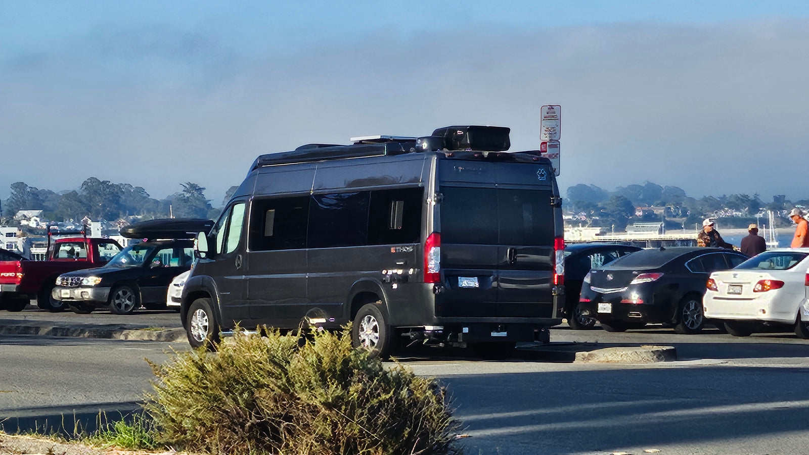 A dark grey camper van, like an RV rental, with roof storage, parked in a sunny lot with other vehicles and people.