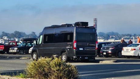 A dark grey camper van, like an RV rental, with roof storage, parked in a sunny lot with other vehicles and people.