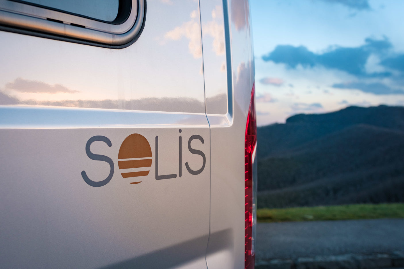 White Solis camper van (an RV rental) shown from the side with its logo, set against a blurry mountain landscape.