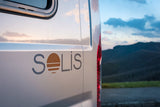White Solis camper van (an RV rental) shown from the side with its logo, set against a blurry mountain landscape.