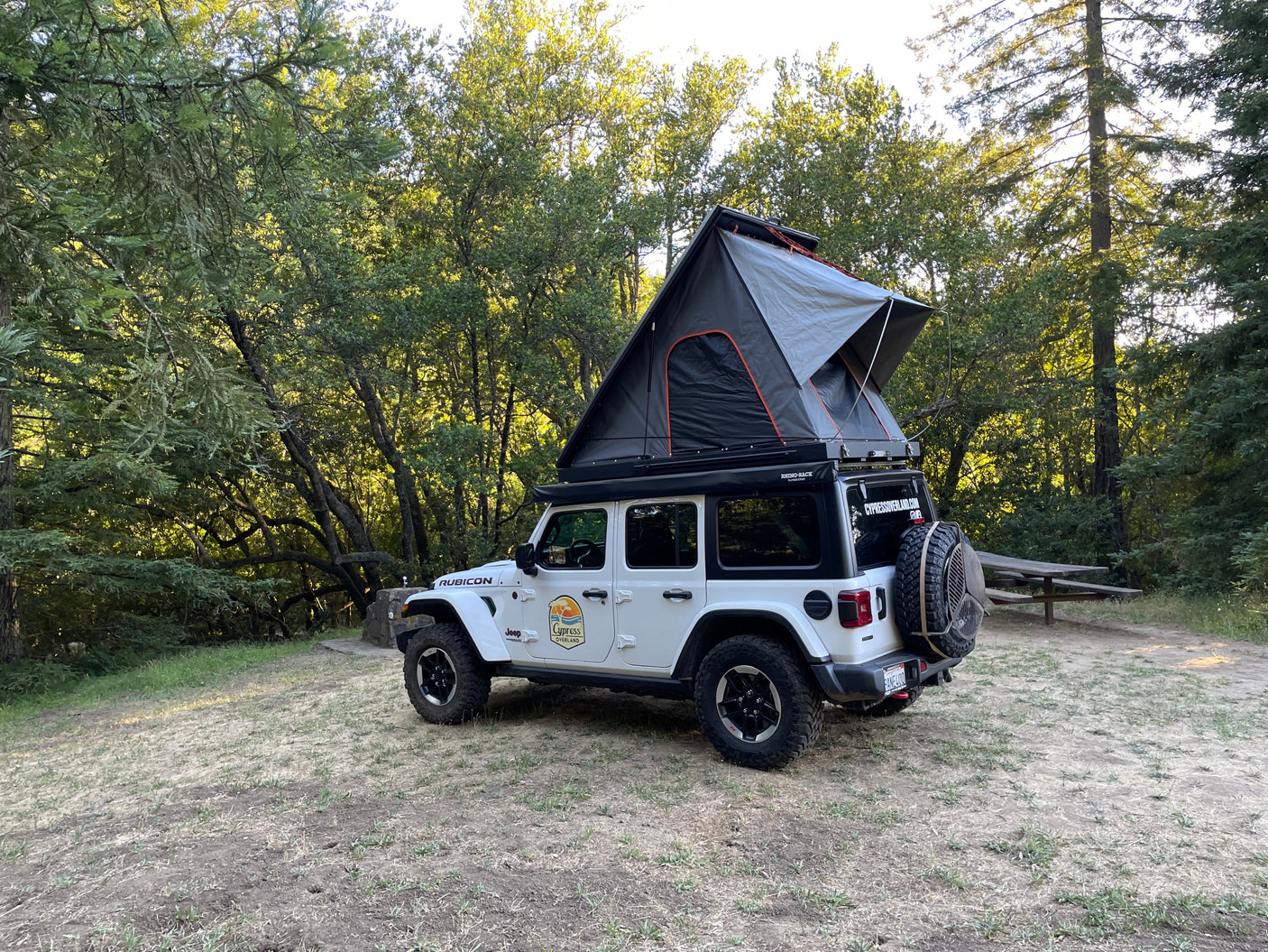 This white Jeep Wrangler with its deployed rooftop tent is parked in a sunny forest campsite, highlighting a unique rv rental option.