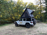 This white Jeep Wrangler with its deployed rooftop tent is parked in a sunny forest campsite, highlighting a unique rv rental option.