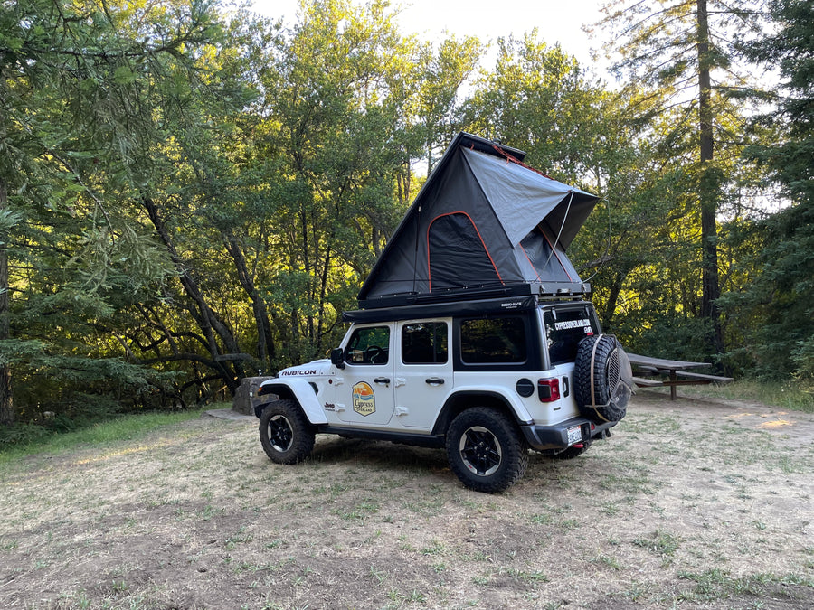 This white Jeep Wrangler with its deployed rooftop tent is parked in a sunny forest campsite, highlighting a unique rv rental option.