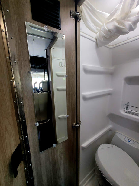 Compact RV rental bathroom with a white shower enclosure, toilet, and a mirror reflecting the cabin interior.