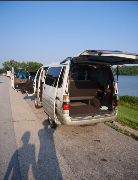 White camper van, resembling an RV rental, parked lakeside with open doors. Shadows of two people visible on the road.