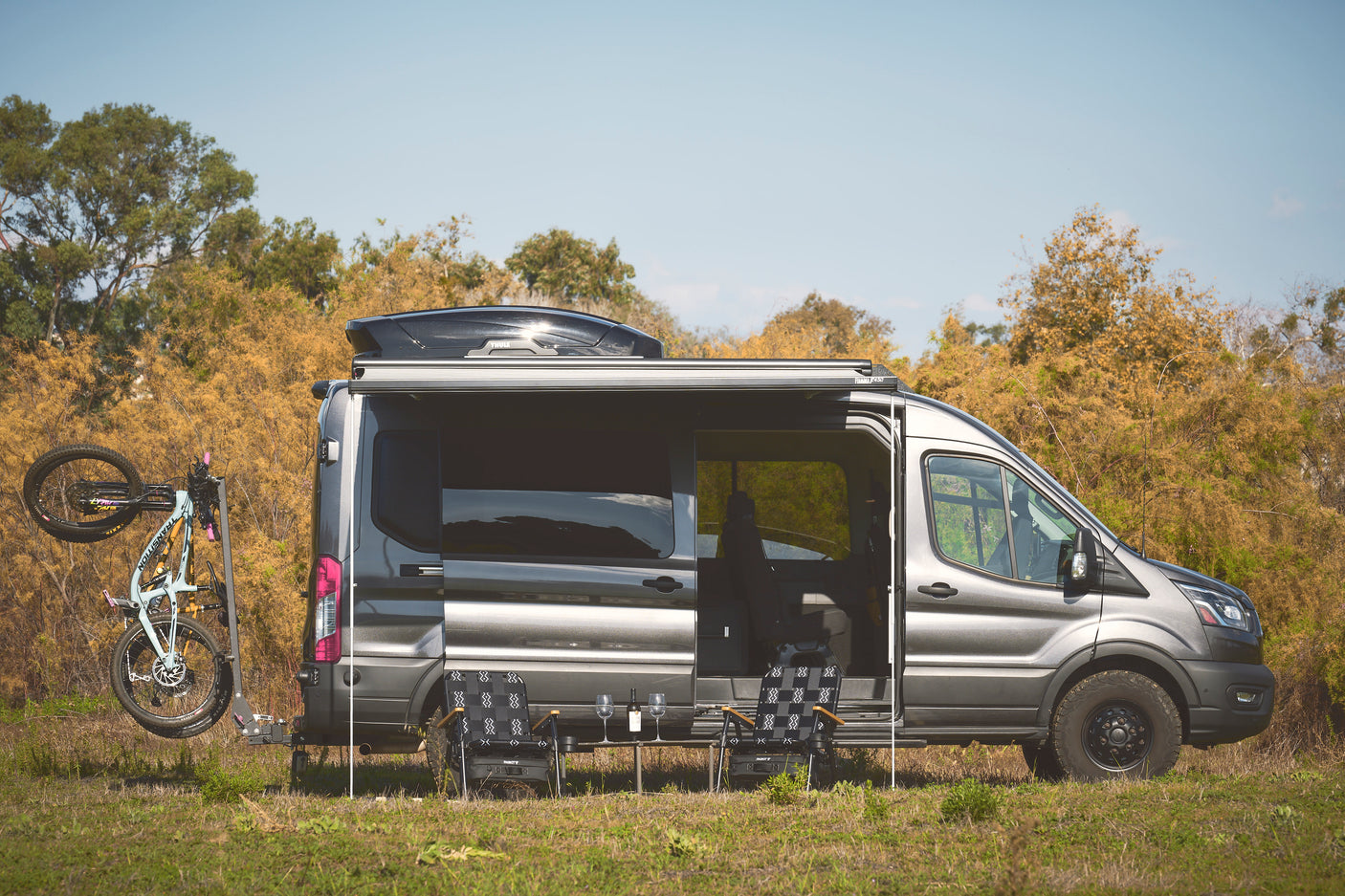Gray RV rental camper van with a bike, extended awning, and outdoor seating setup, parked in a grassy field.