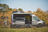 Gray RV rental camper van with a bike, extended awning, and outdoor seating setup, parked in a grassy field.